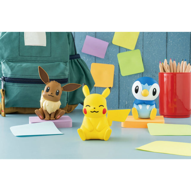 A model kit figure of Pikachu, the Electric-type Pokémon, sitting down with a cheerful expression. Its eyes are closed, and its mouth forms a small, happy smile. The figure is bright yellow with red cheeks, large pointy ears with black tips, and a distinctive lightning bolt-shaped tail extending to the side. Its short arms rest on its round belly, and its feet are positioned slightly apart. The background has a school scene behind.