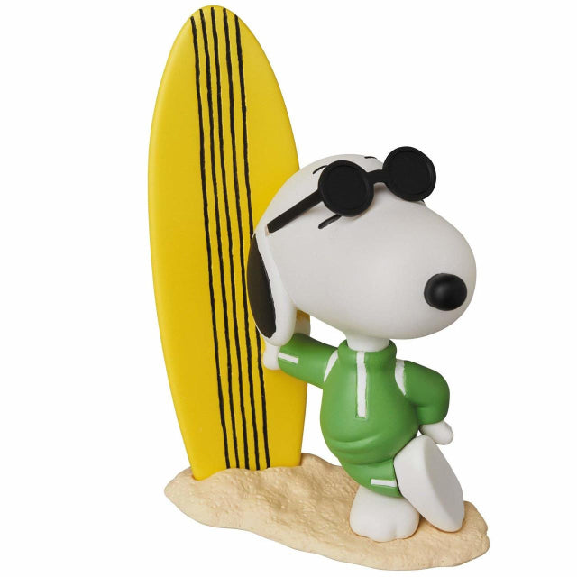 A stylish Snoopy figure from the Peanuts series, dressed as Joe Cool. Snoopy is leaning casually against a tall yellow surfboard with black stripes, wearing a green zip-up wetsuit and black sunglasses. His relaxed pose and confident expression capture the laid-back surfer vibe. The figure is set on a sandy base, evoking a classic beach scene.