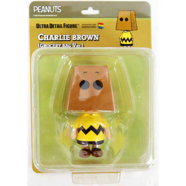 A unique Charlie Brown figure from the Peanuts series, wearing his classic yellow shirt with a black zigzag stripe, black shorts, and brown shoes. Instead of his usual expression, Charlie Brown has a large brown paper bag over his head, with two round cut outs for eyes and simple drawn-on eyebrows, giving him a humorous and slightly shy appearance.