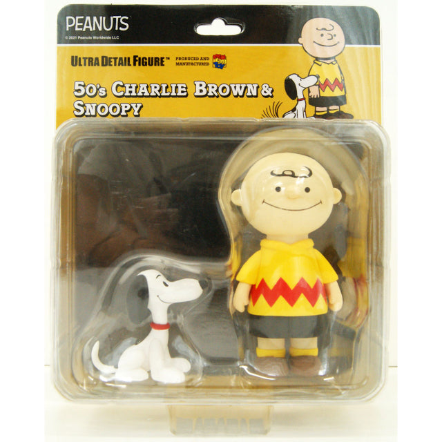 A collectible figure set featuring Charlie Brown and Snoopy from the Peanuts series. Charlie Brown is depicted in a vintage 1950s style, wearing his signature yellow shirt with a red zigzag pattern, black shorts, yellow socks, and brown shoes. He has a round head with a small tuft of hair and a simple, smiling expression. Snoopy, sitting beside him, has a classic cartoonish design with a white body, black ears, a black nose, and a red collar. Both figures have a smooth, minimalistic design reminiscent of ea