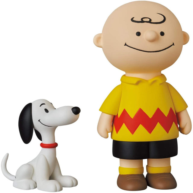 A collectible figure set featuring Charlie Brown and Snoopy from the Peanuts series. Charlie Brown is depicted in a vintage 1950s style, wearing his signature yellow shirt with a red zigzag pattern, black shorts, yellow socks, and brown shoes. He has a round head with a small tuft of hair and a simple, smiling expression. Snoopy, sitting beside him, has a classic cartoonish design with a white body, black ears, a black nose, and a red collar. Both figures have a smooth, minimalistic design reminiscent of ea