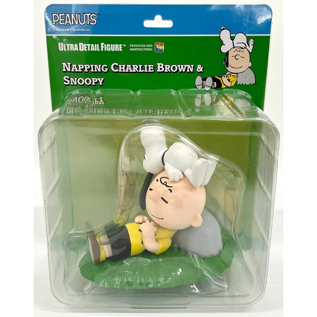 A figure of Charlie Brown and Snoopy from the Peanuts series. Charlie Brown is sitting on the grass, leaning against a gray rock with a relaxed smile, eyes closed, and hands resting on his chest. He wears a yellow shirt with a black zigzag pattern, black shorts, white socks, and brown shoes. Snoopy, the white beagle with black ears, is lying upside down on Charlie Brown’s head, his paws stretched out and his black nose pointing downward. The figure is designed in a soft, cartoonish style with a smooth finis